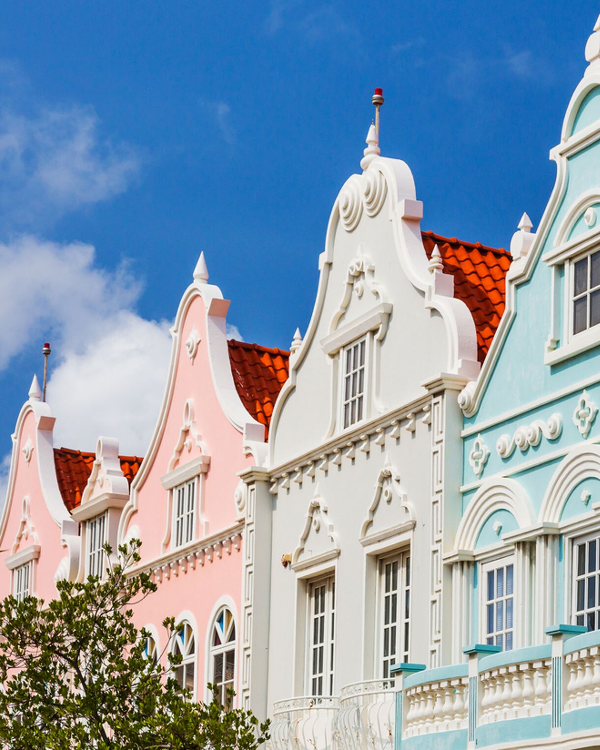A row of colorful Dutch colonial buildings in Oranjestad, Aruba, featuring intricate white trim, arched windows and terracotta-tiled roofs against a bright blue sky.