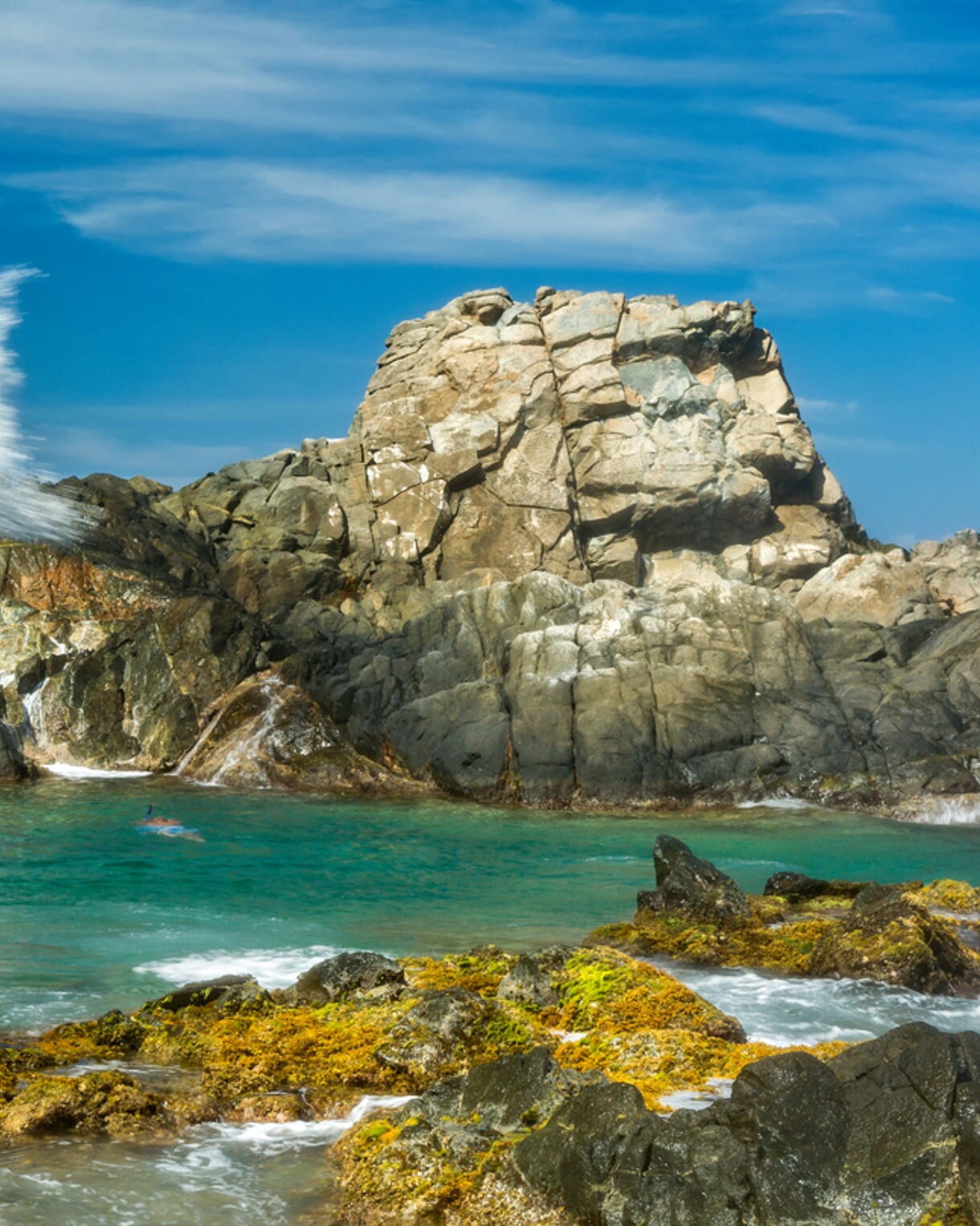 A secluded natural pool in Aruba surrounded by rugged volcanic rocks, with waves crashing dramatically against the shoreline and a swimmer floating in the turquoise water.