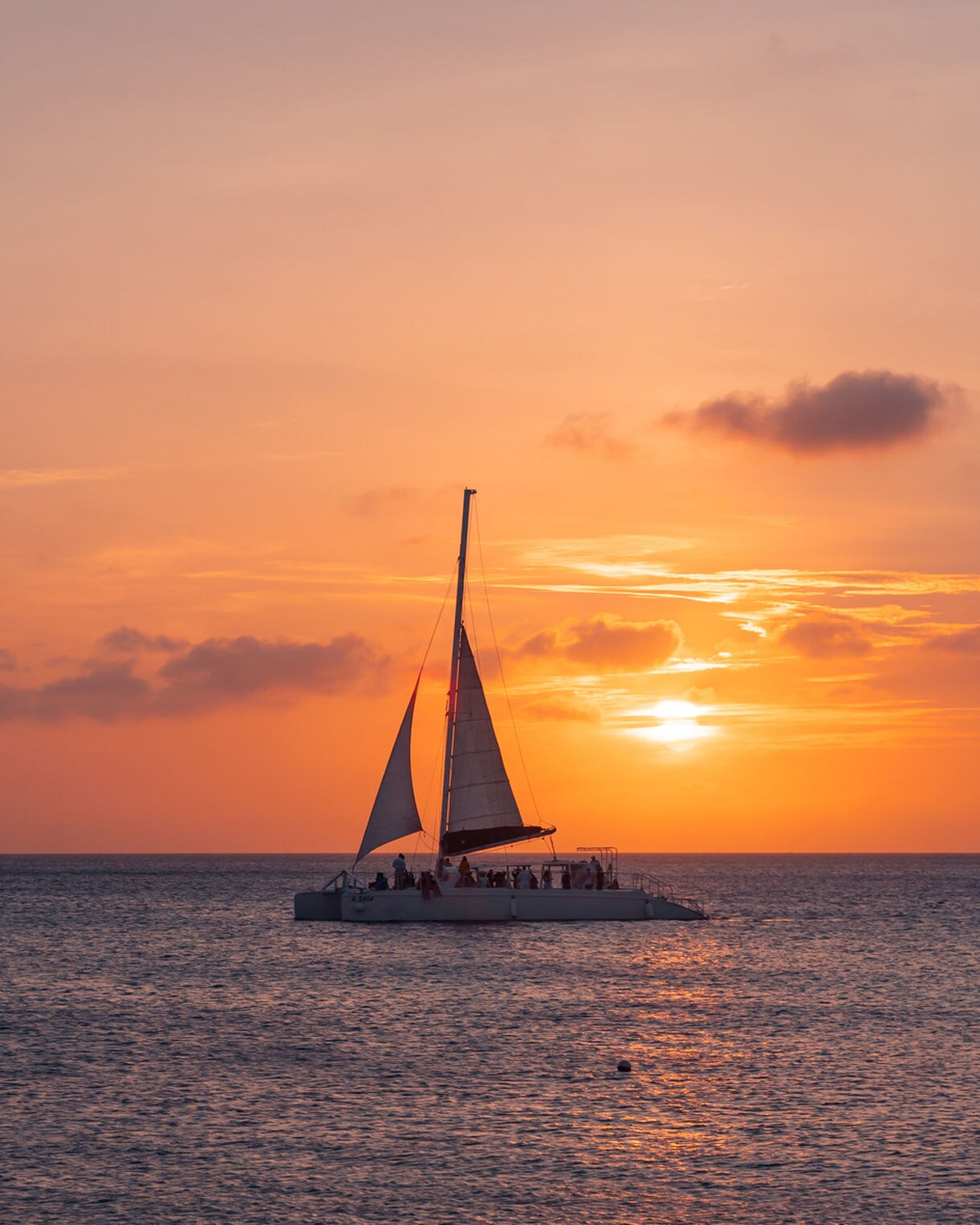 A catamaran with white sails glides across the ocean at sunset, silhouetted against a sky painted in warm orange and pink tones.