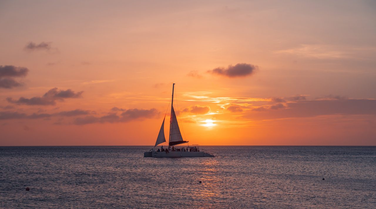 A catamaran with white sails glides across the ocean at sunset, silhouetted against a sky painted in warm orange and pink tones.
