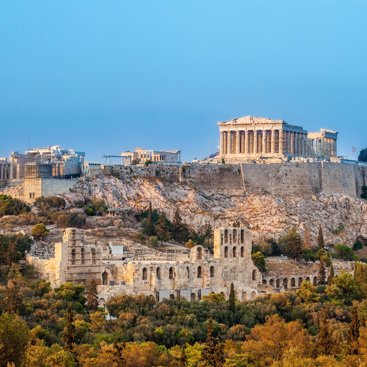 The Acropolis of Athens with the Parthenon and ancient ruins lit by evening light against a clear blue sky.