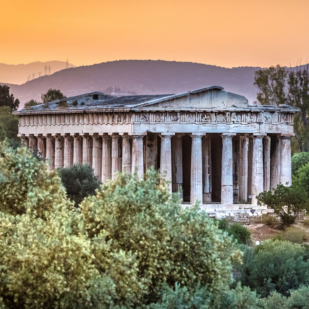 The Temple of Hephaestus framed by trees at sunset with soft orange skies and hills in the background.