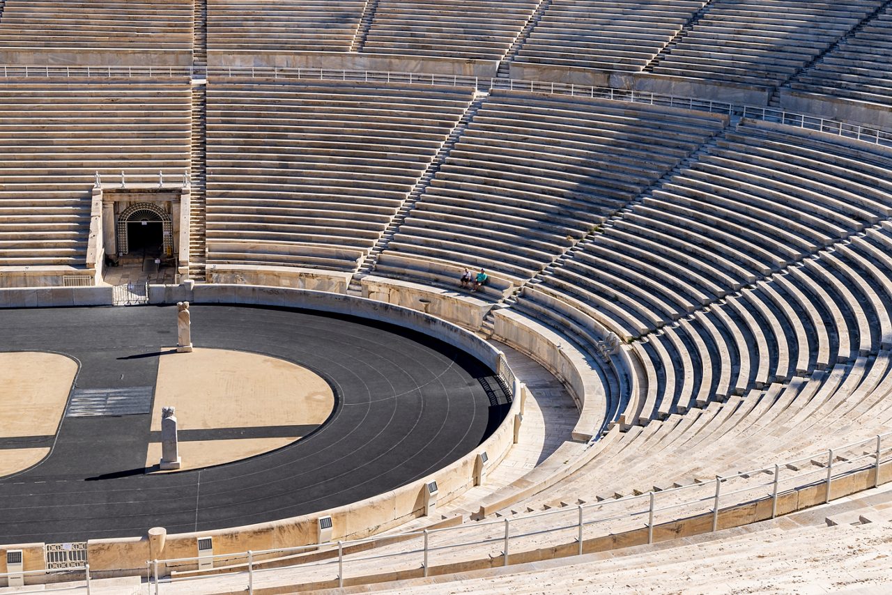 The Panathenaic Stadium in Athens with tiered marble seating curving around a long track, surrounded by trees and city buildings under a partly cloudy sky