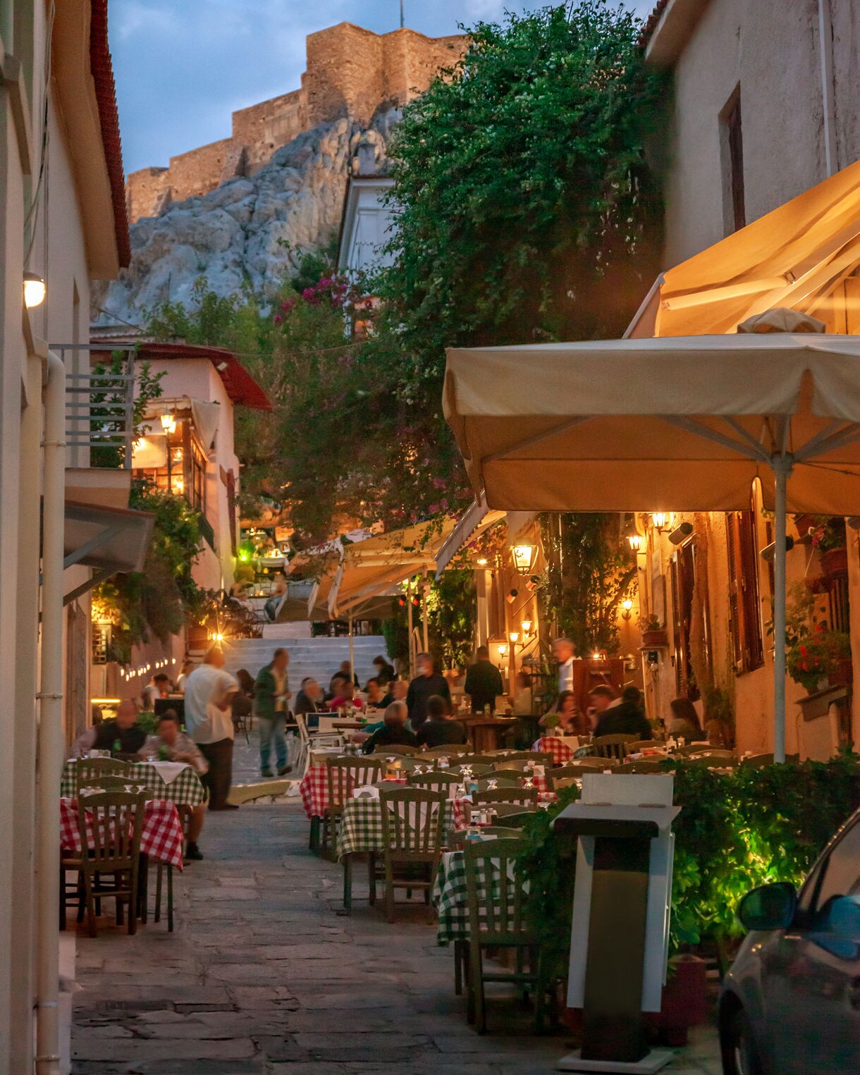Evening view of a lively Athens street with outdoor tavernas, colorful tablecloths, and the Acropolis lit in the background.