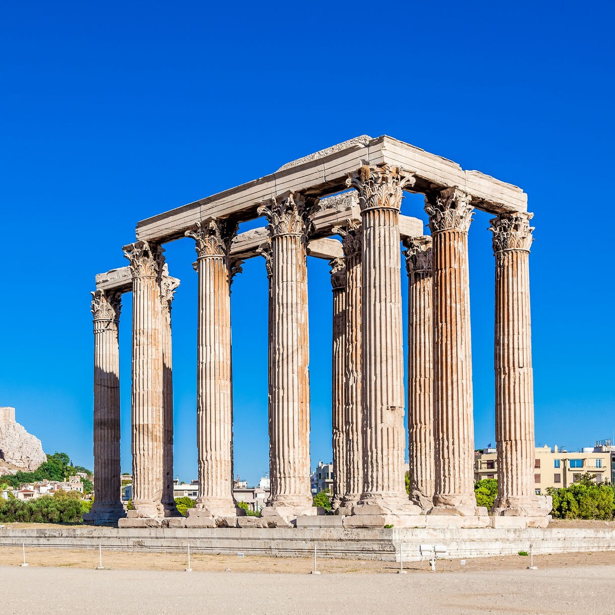 Ancient stone columns of the Temple of Olympian Zeus standing tall against a vivid blue sky with the Acropolis in the background.