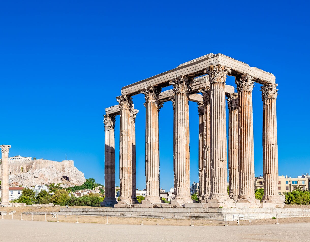 The Temple of Olympian Zeus in Athens with massive Corinthian columns standing under a clear blue sky and the Acropolis visible in the background.