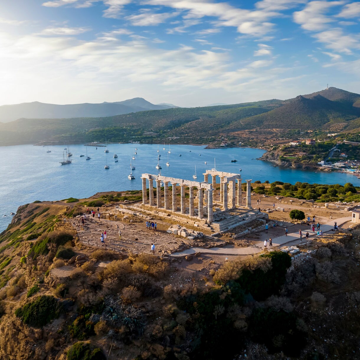 Aerial view of the Temple of Poseidon perched on a cliff above the sea with sailboats and green hills in the background.