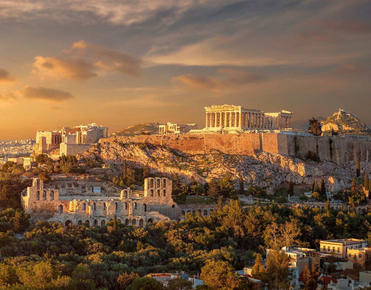 The Acropolis of Athens at sunset with the Parthenon and ancient ruins lit by warm golden light above the city.