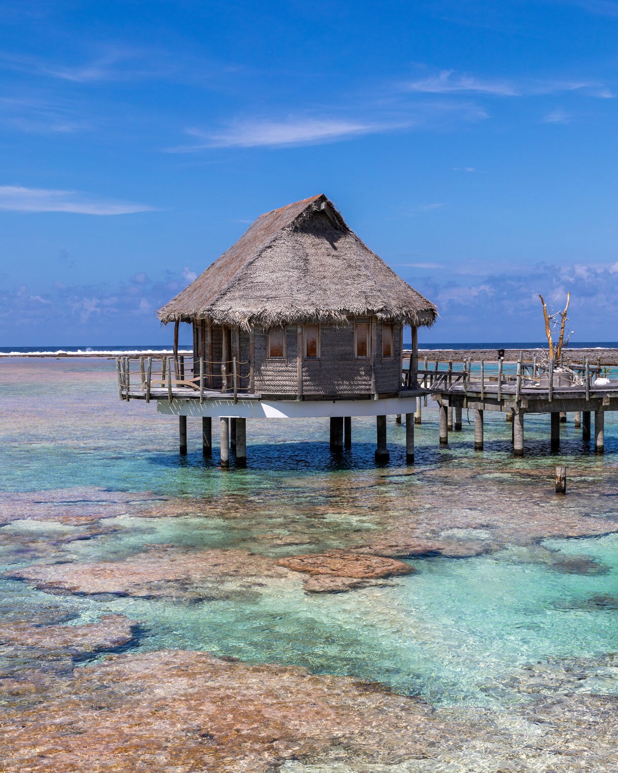 Thatched-roof overwater bungalow on stilts above turquoise lagoon with coral reefs visible below.