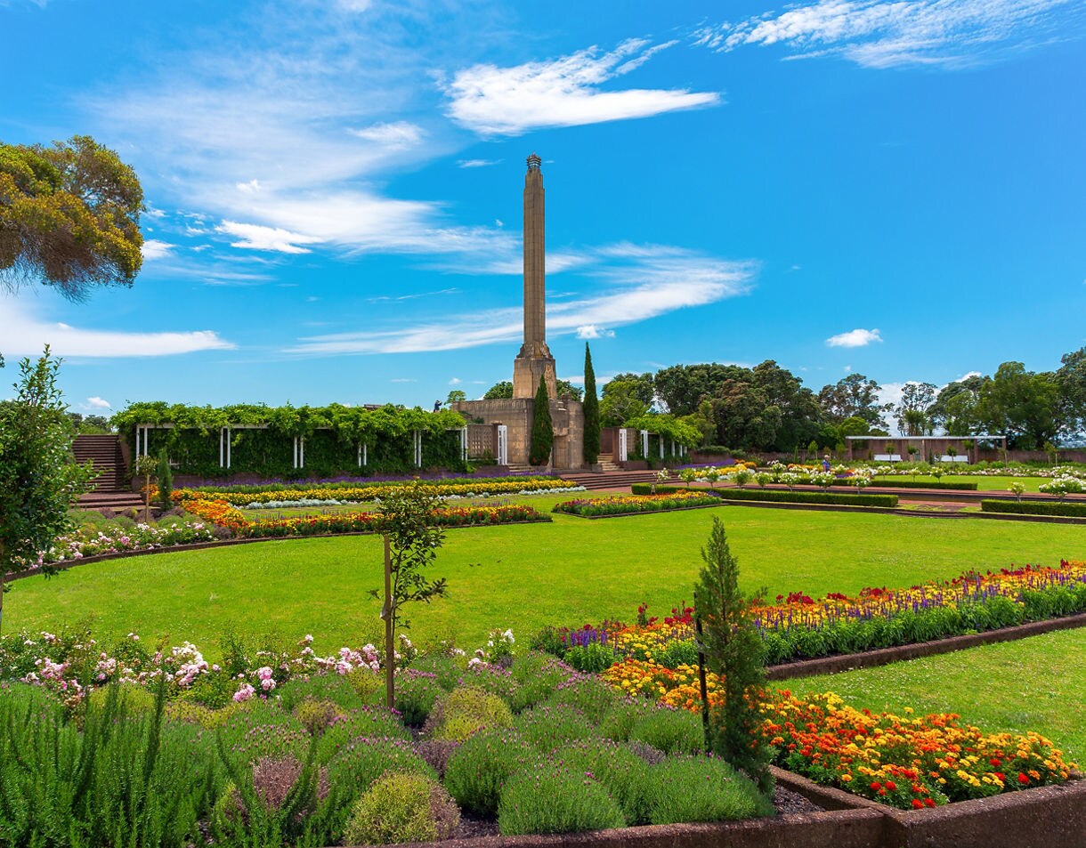 A large landscaped garden with colorful flowerbeds and neatly trimmed lawns surrounding a tall memorial obelisk at Bastion Point, with trees and a bright blue sky in the background.