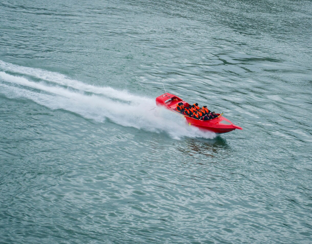 A bright red jet boat with passengers in orange life jackets speeding across blue-green harbor water, leaving a white spray trail behind.