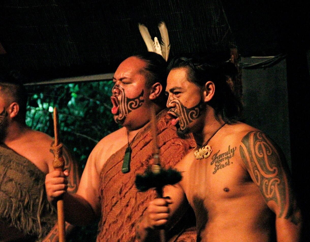 Three Māori performers in traditional attire with facial moko, holding carved wooden implements, chanting with expressive intensity against a dark, natural backdrop.