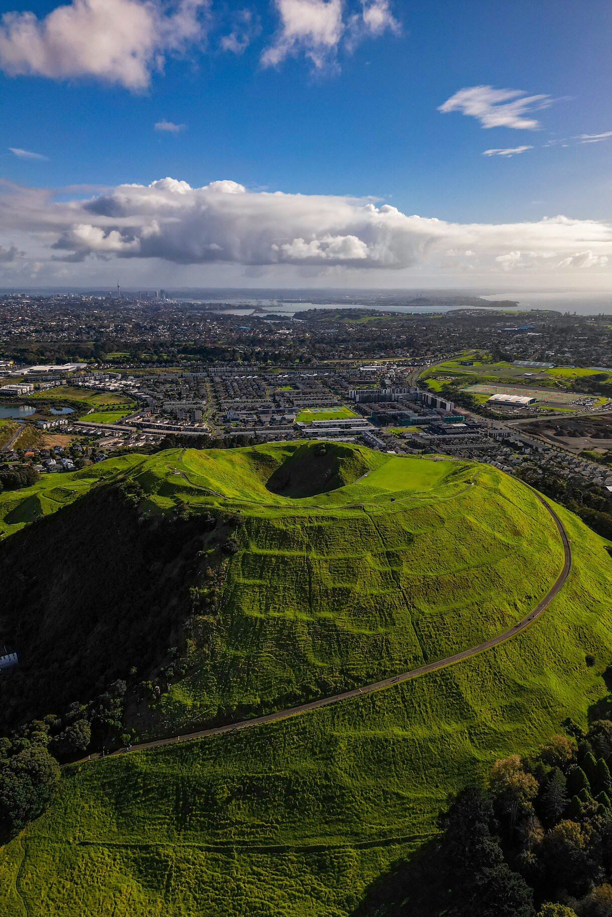 Aerial view of a grassy volcanic crater surrounded by city neighborhoods, with water and distant skyline under a bright, partly cloudy sky.