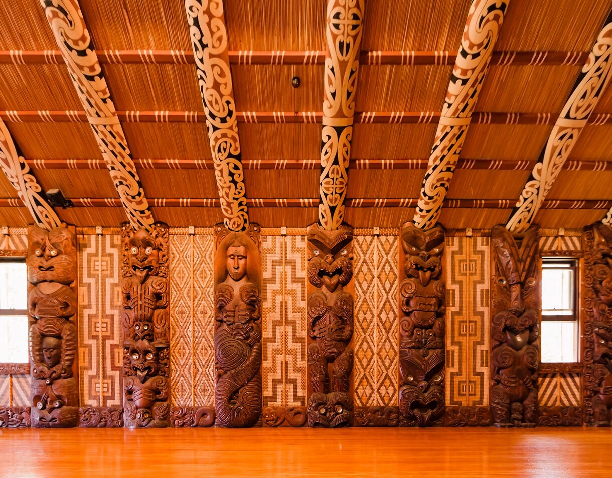 Interior of a Māori wharenui with ornate wooden carvings and patterned panels, warm light across polished wood floor.