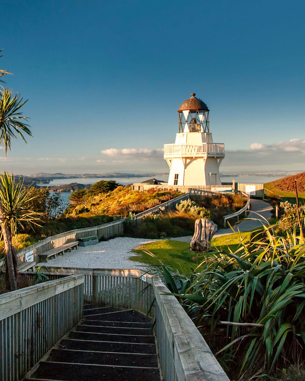 View of Manukau Heads Lighthouse in Auckland, New Zealand, perched on a cliff overlooking the ocean, with waves crashing against the rocky shore and a clear blue sky above.