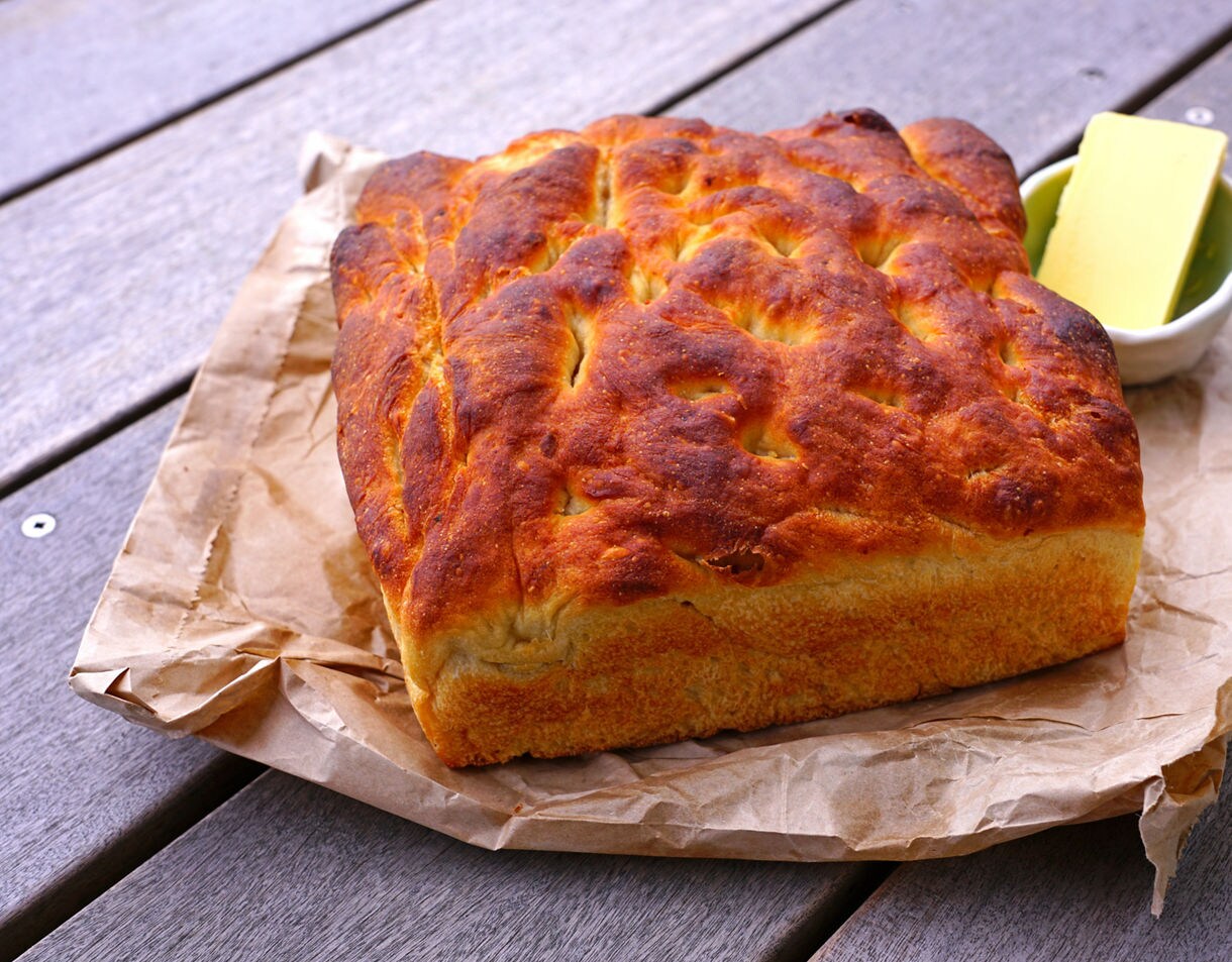 Close-up of a golden, rustic loaf of rewena bread resting on brown paper, with a small dish of butter beside it on a wooden table.