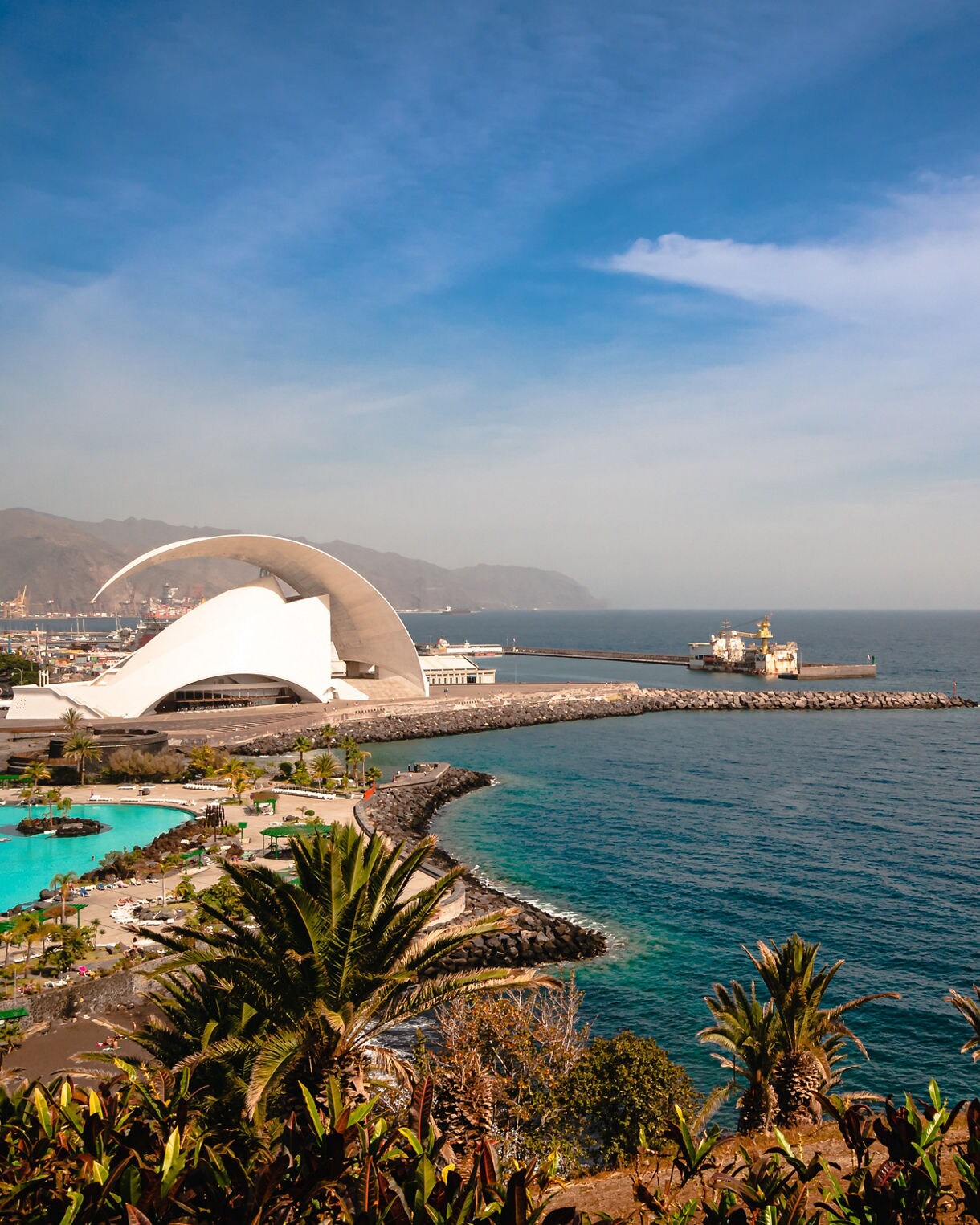 White curved structure of the Auditorio de Tenerife by the ocean, with turquoise pools and palm trees in the foreground.