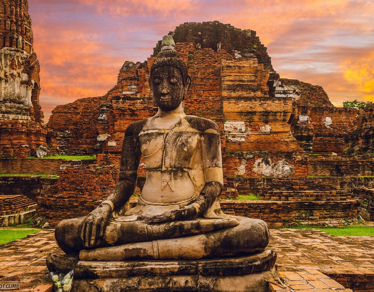 Weathered stone Buddha statue seated in front of red brick ruins and temple walls, glowing under an orange and pink sunset sky.