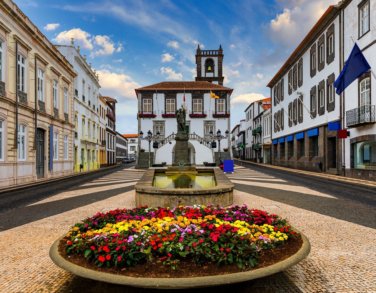 A colorful flowerbed and fountain in front of Angra do Heroísmo’s town hall, surrounded by whitewashed buildings and patterned cobblestone streets.