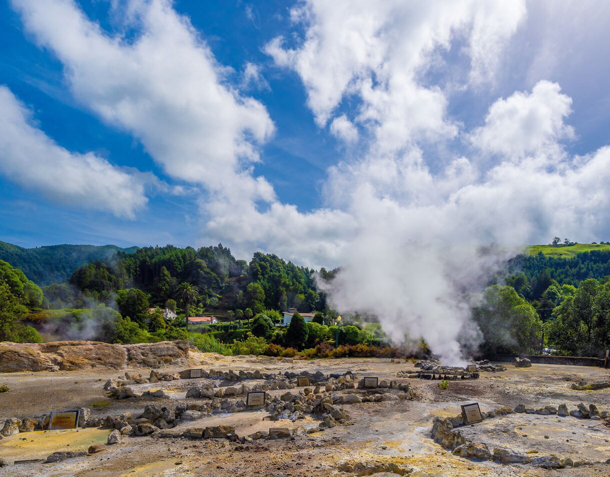 Geothermal vents releasing steam amid rocky terrain in Furnas Valley, surrounded by lush green hills under a bright blue sky.