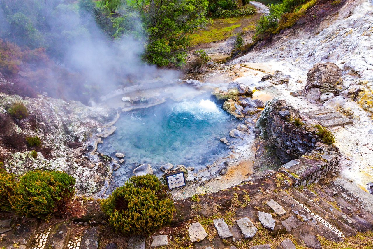 A steaming blue geothermal pool surrounded by rocky terrain and greenery in Furnas Valley on São Miguel Island.