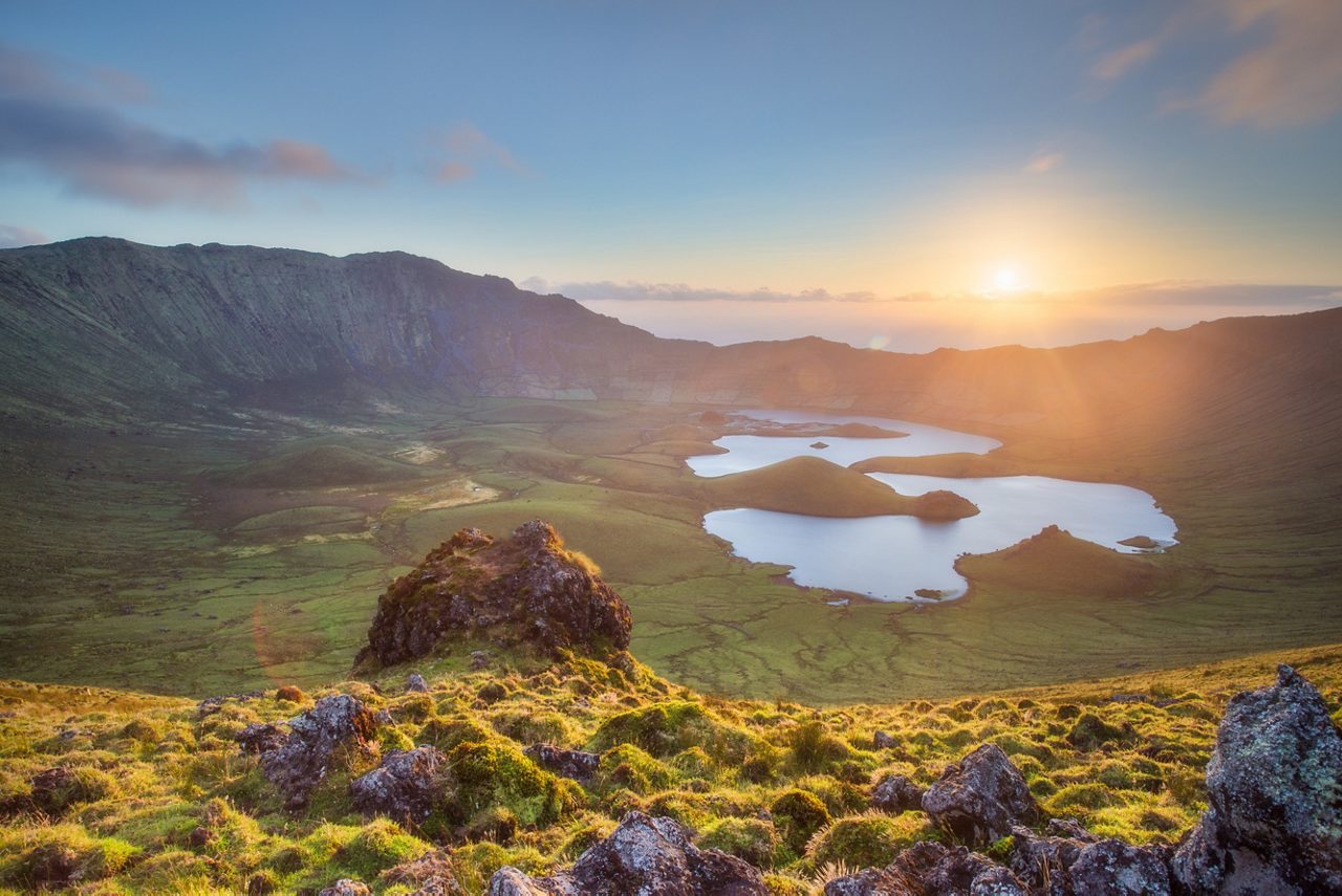 A scenic landscape of volcanic crater lakes in the Azores, Portugal, with rolling green hills and a glowing sunset casting light over the peaceful valley.