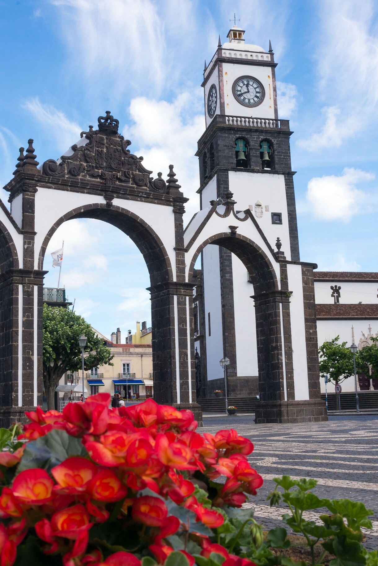 The Portas da Cidade arches and clock tower in Ponta Delgada framed by red flowers and patterned stone pavement under a blue sky.