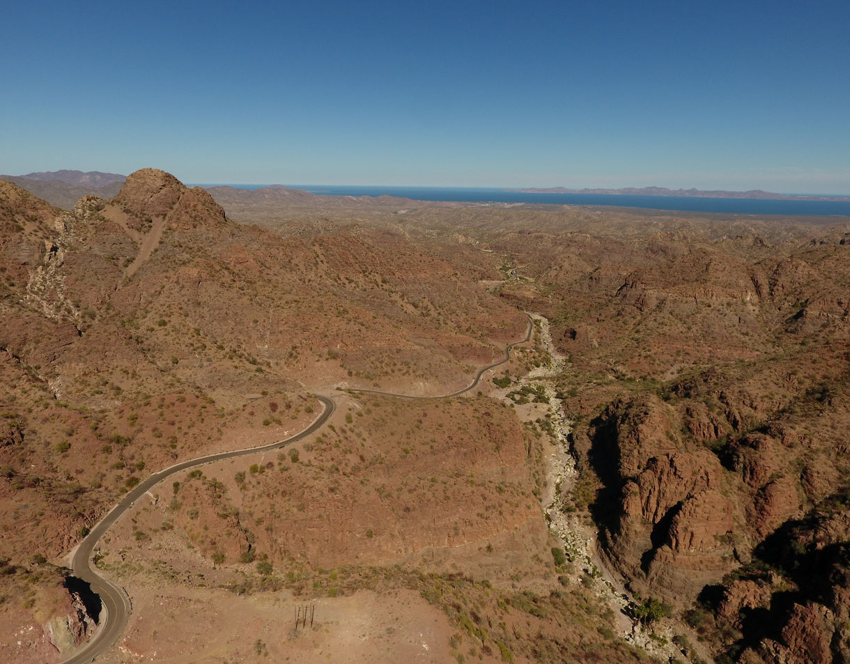 Aerial view of Baja California desert with winding roads weaving through arid, rocky mountains leading toward the distant sea.