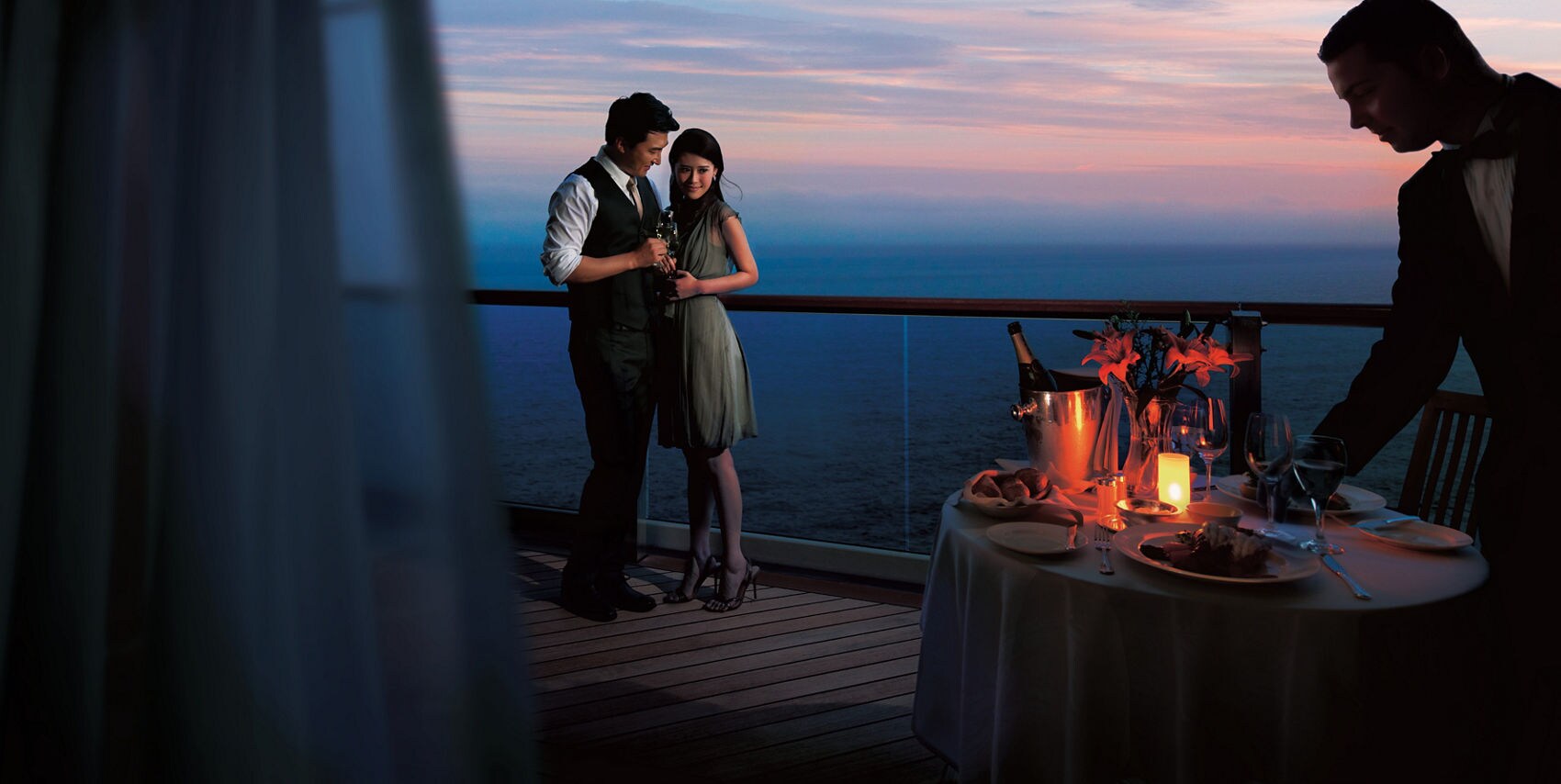 Room steward sets up dinner table while couple embraces on stateroom balcony