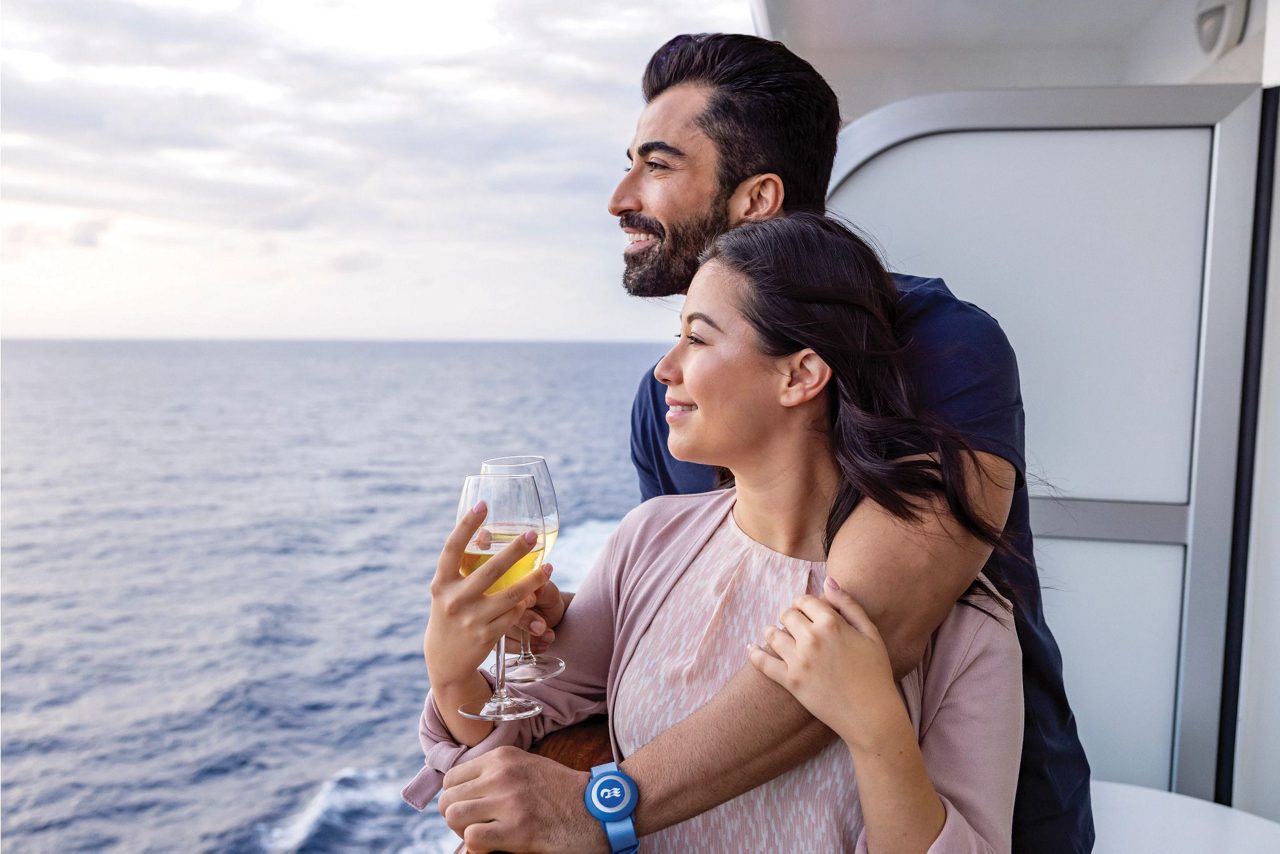 A couple standing on a cruise ship balcony, gazing at the ocean horizon with smiles. The woman holds a glass of wine, and both appear relaxed, enjoying a serene moment together on the open sea.