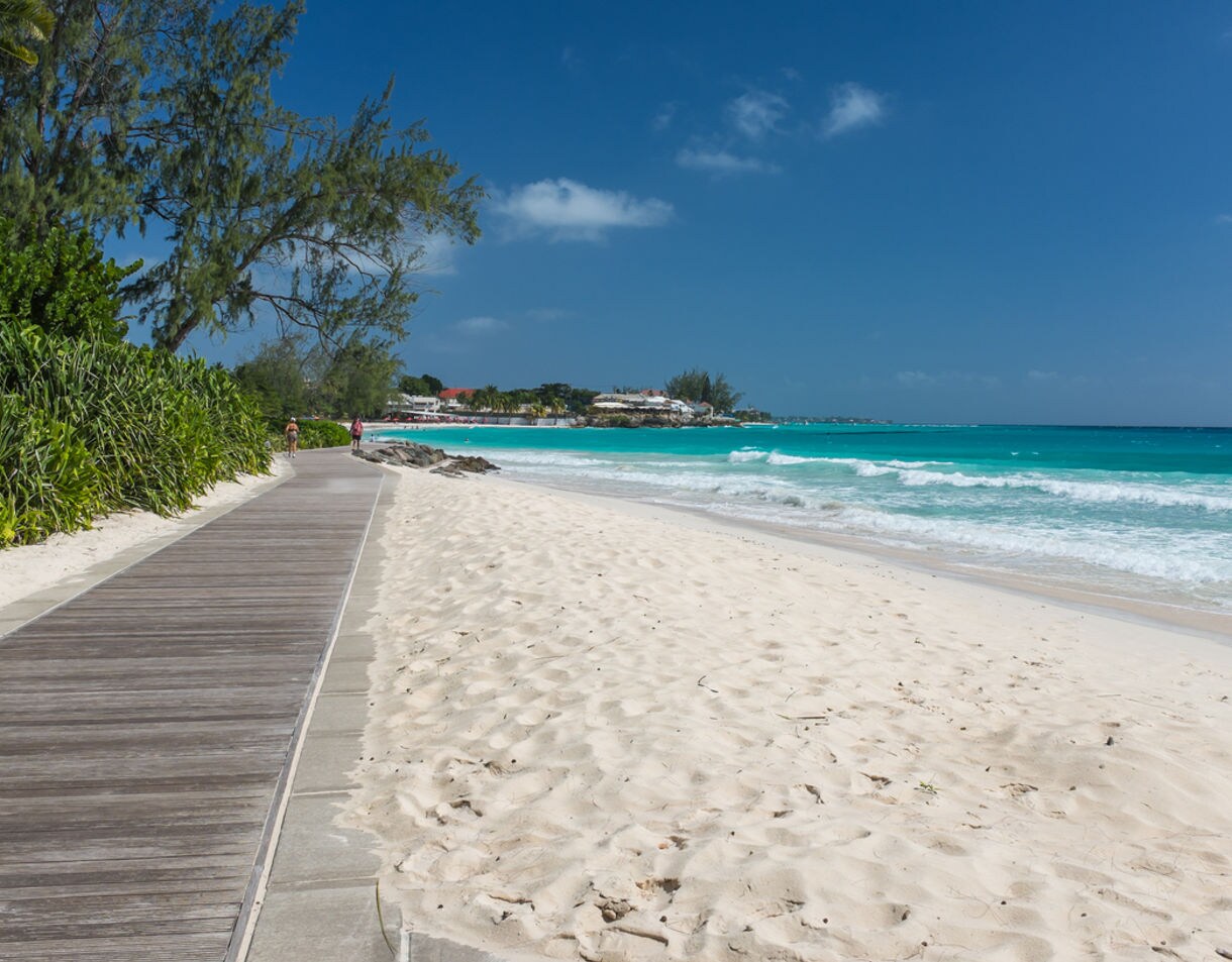 Coastal boardwalk beside Accra Beach in Barbados with soft white sand, turquoise waves and tropical greenery under a clear blue sky.