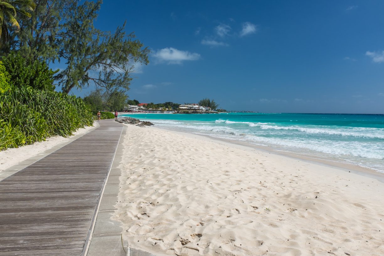 Coastal boardwalk beside Accra Beach in Barbados with soft white sand, turquoise waves and tropical greenery under a clear blue sky.