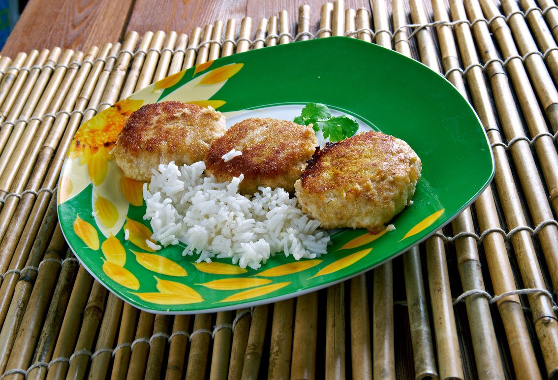 Close-up of a plate with three golden-brown fish cakes and a portion of white rice, garnished with a sprig of parsley, placed on a bamboo mat.