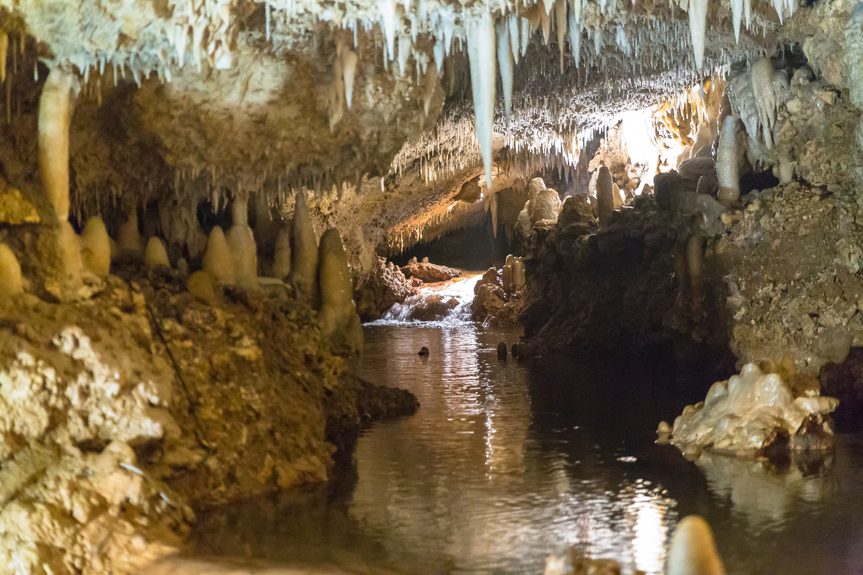  An illuminated passage inside Harrison’s Cave in Barbados featuring a reflective underground stream, towering stalagmites and sharp stalactites hanging from the ceiling.
