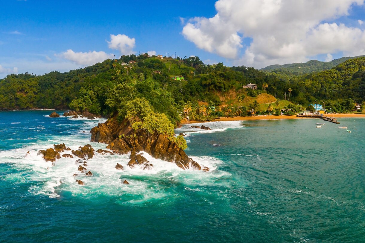 Panoramic aerial view of a tropical coastline with vibrant turquoise water, rocky outcrops, dense green hills and a small village with boats anchored near a sandy beach.