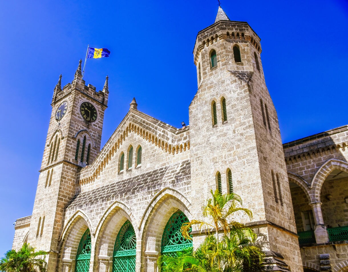 Stone Gothic-style Parliament Buildings in Bridgetown, Barbados with arched green doorways, clock towers and the national flag flying under a clear blue sky