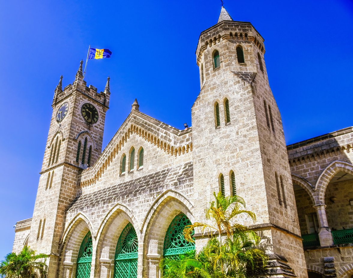 Stone Gothic-style Parliament Buildings in Bridgetown, Barbados with arched green doorways, clock towers and the national flag flying under a clear blue sky