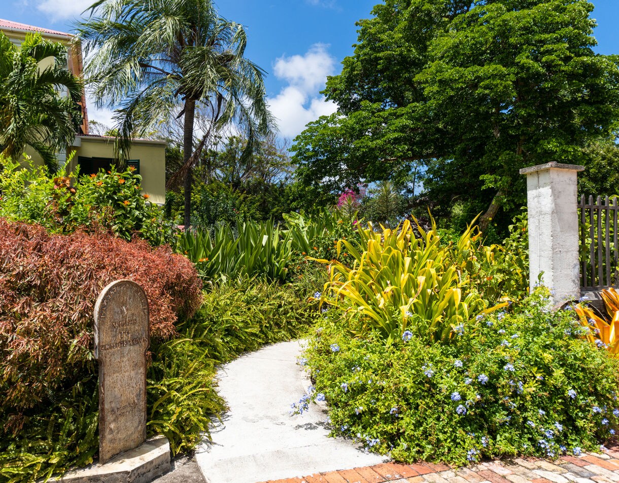 Bright garden path surrounded by colorful tropical plants and palm trees at Sunbury Plantation House in Barbados, with a stone marker and a light green building partially visible.