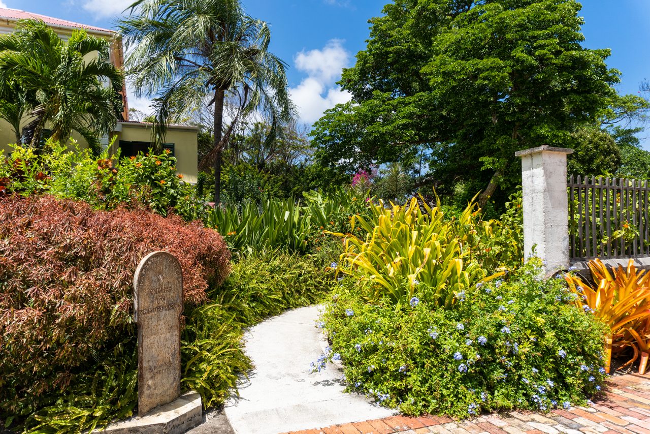 Bright garden path surrounded by colorful tropical plants and palm trees at Sunbury Plantation House in Barbados, with a stone marker and a light green building partially visible.