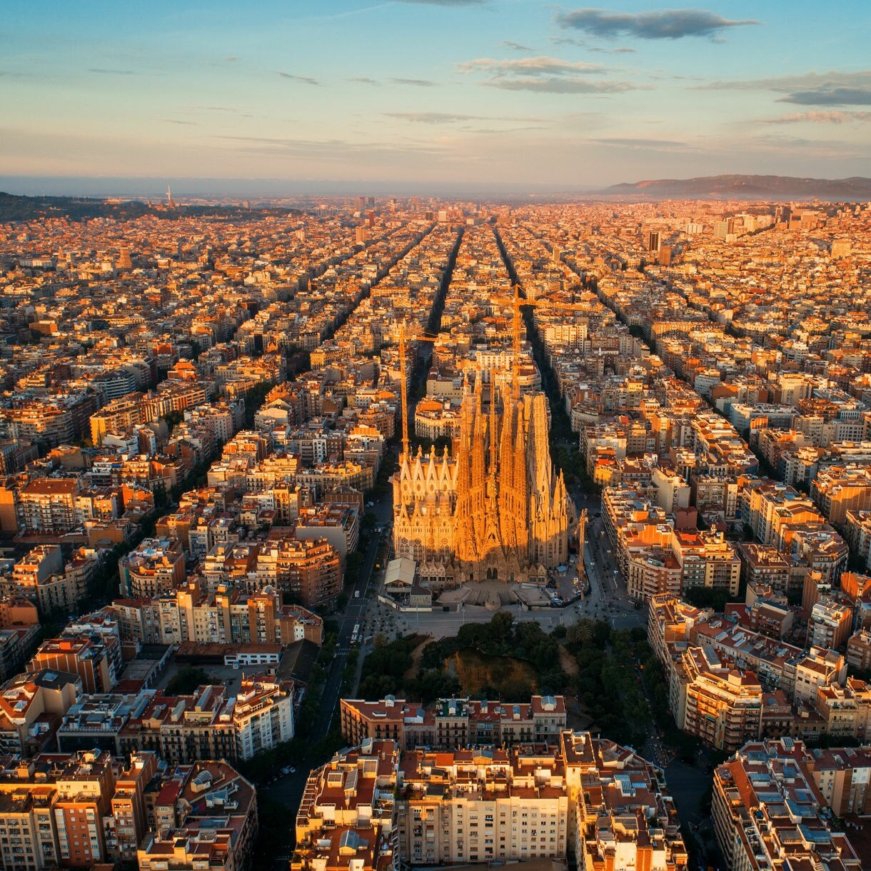 Sweeping aerial view of Barcelona bathed in warm sunset light, with the Sagrada Família's soaring spires rising from the center of the city's vast geometric grid of octagonal blocks stretching to the hills and sea on the horizon.