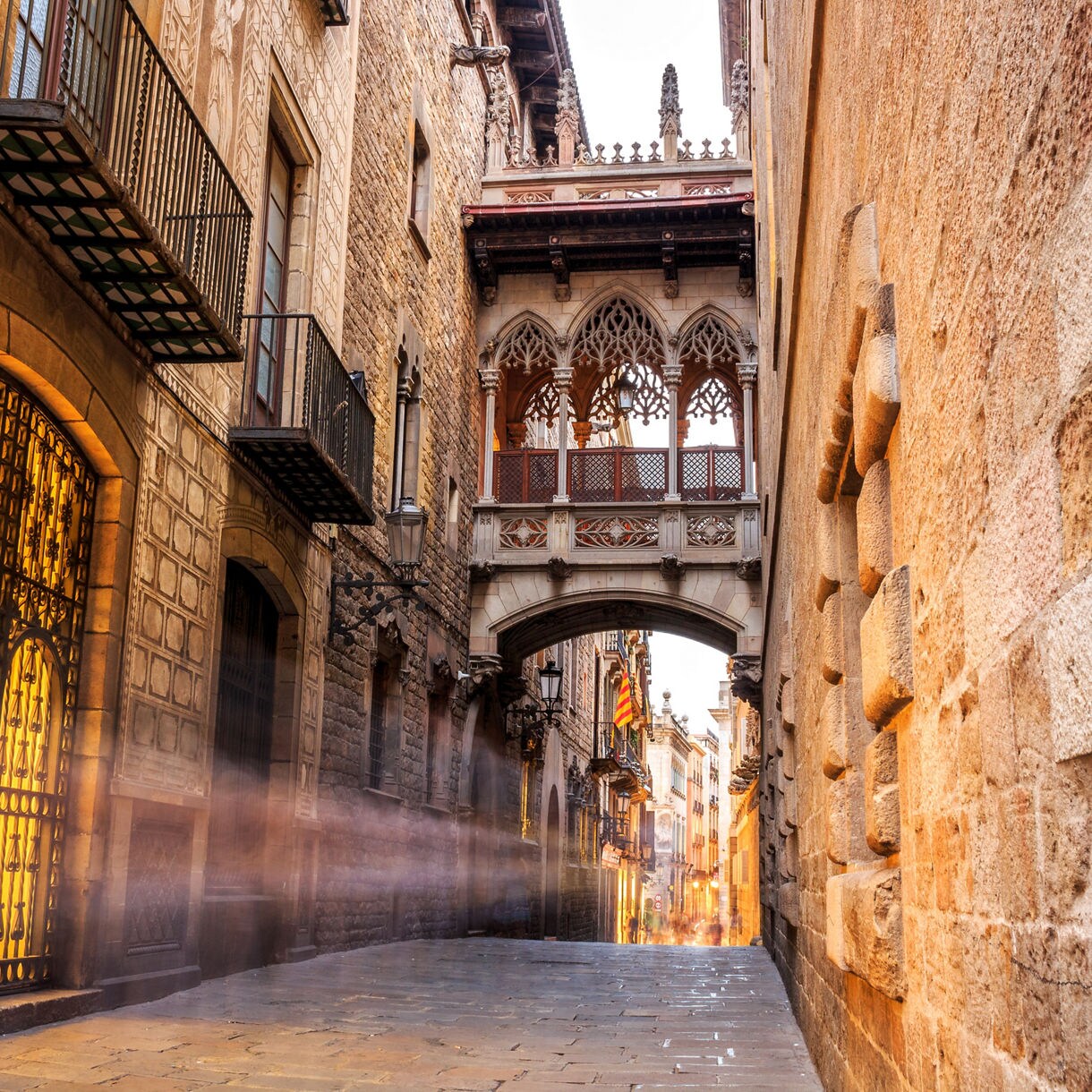 Narrow cobblestone street in Barcelona’s Gothic Quarter featuring the iconic neo-Gothic Pont del Bisbe bridge, surrounded by ornate stone buildings with balconies and warm interior lighting.
