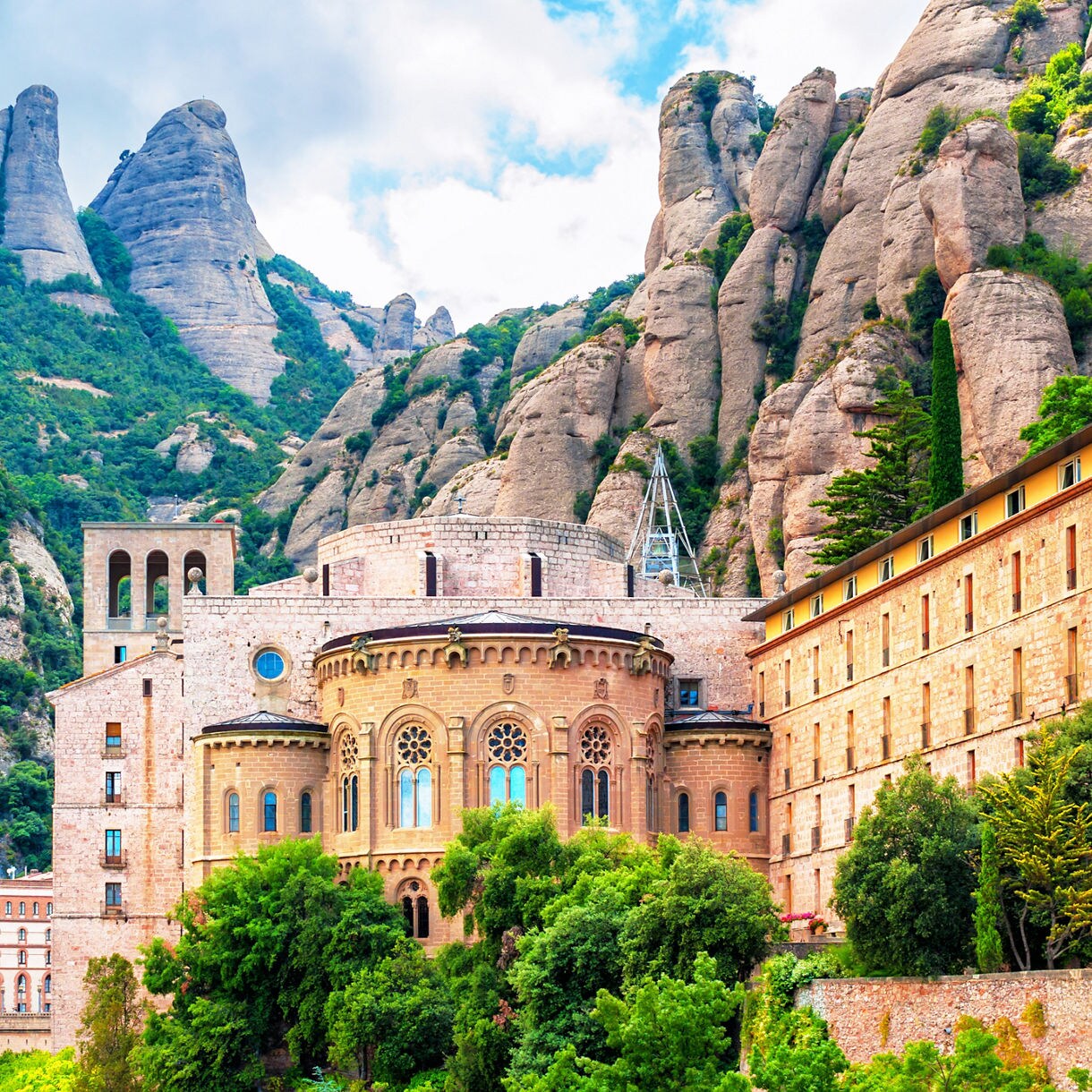 View of the Montserrat Monastery nestled against jagged limestone cliffs in Catalonia, Spain, with Romanesque and Gothic structures surrounded by lush green vegetation and steep rocky mountains.