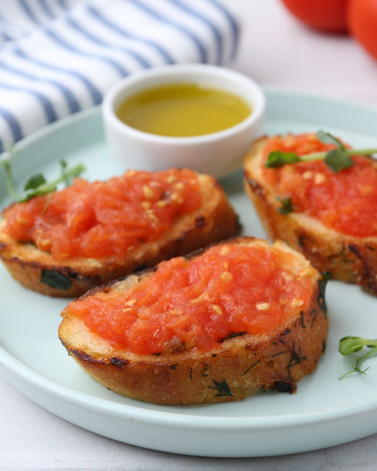 Three slices of toasted bread topped with fresh tomato spread, served with a small dish of olive oil on a light blue plate.