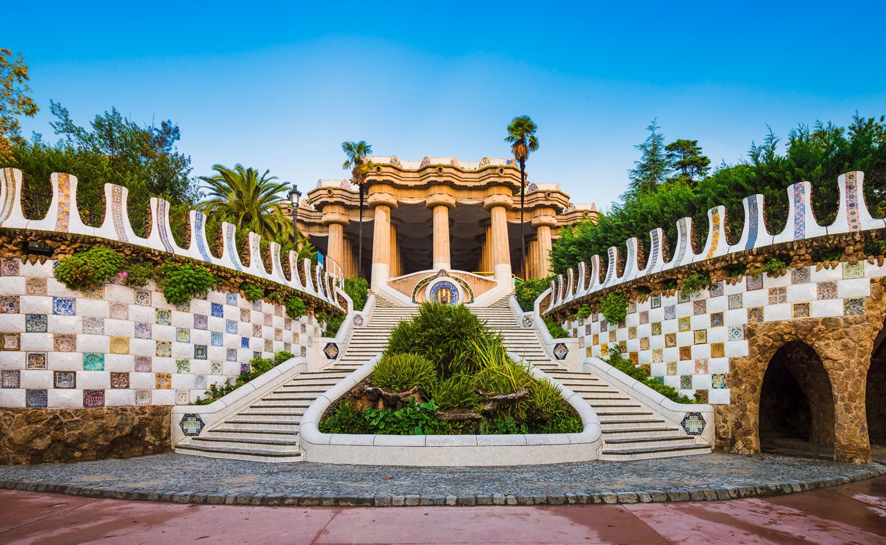 Symmetrical staircase at Park Güell in Barcelona, framed by mosaic-tiled walls and leading up to columned structures surrounded by greenery.
