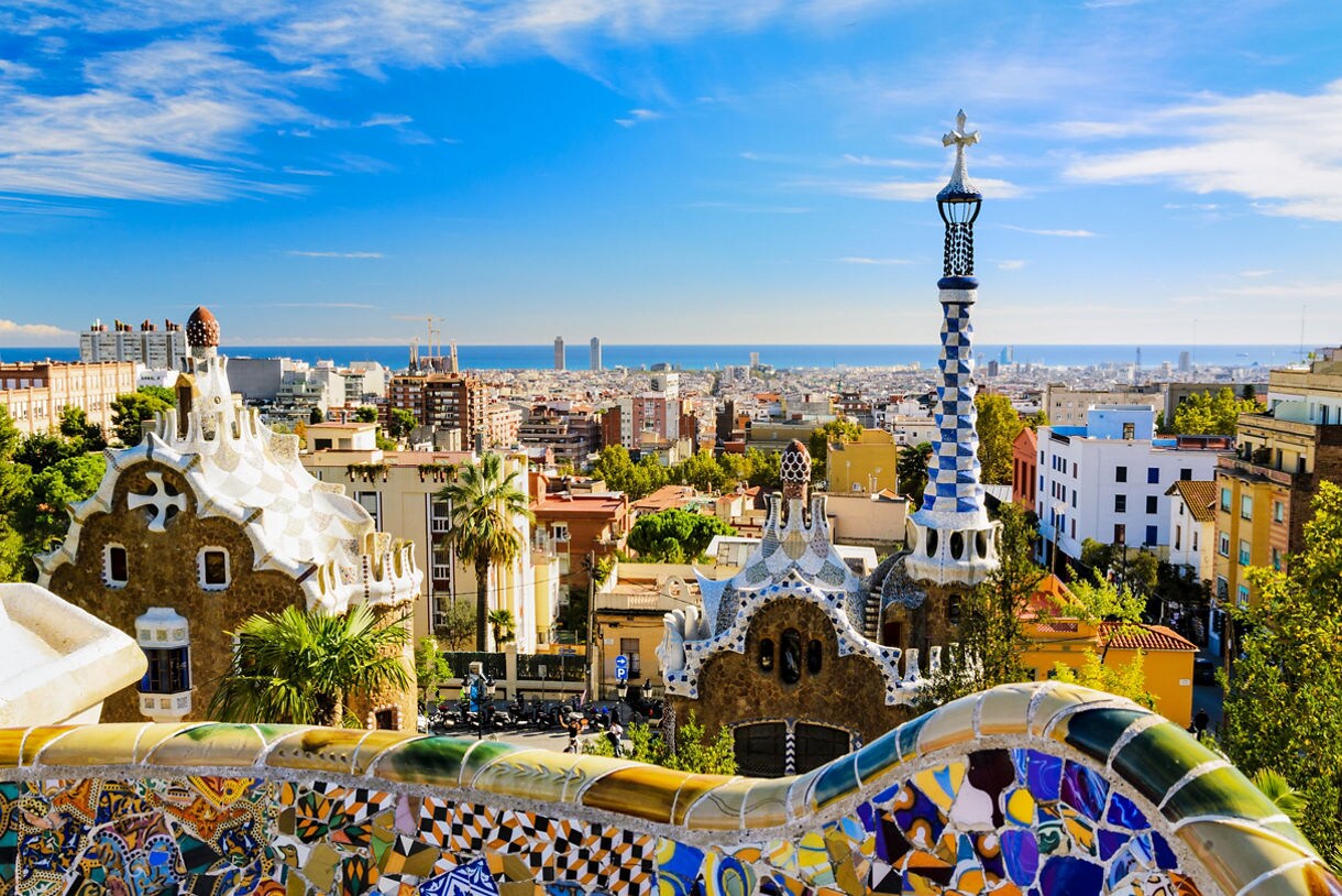 Vibrant mosaic tiles in Park Güell overlooking whimsical Gaudí architecture, with the city of Barcelona and the Mediterranean Sea in the distance.