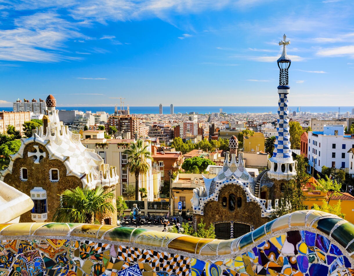 Panoramic view of Barcelona from Park Güell, featuring colorful mosaic tilework, whimsical Gaudí-designed buildings and towers in the foreground, with the city and sea stretching into the distance.