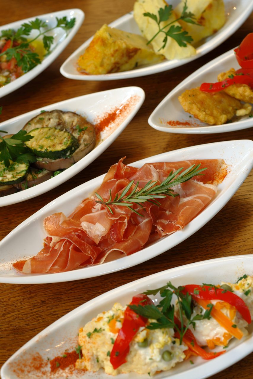 Assorted tapas dishes on small white plates, featuring cured ham, tortilla, grilled vegetables, and creamy salads garnished with herbs and peppers.
