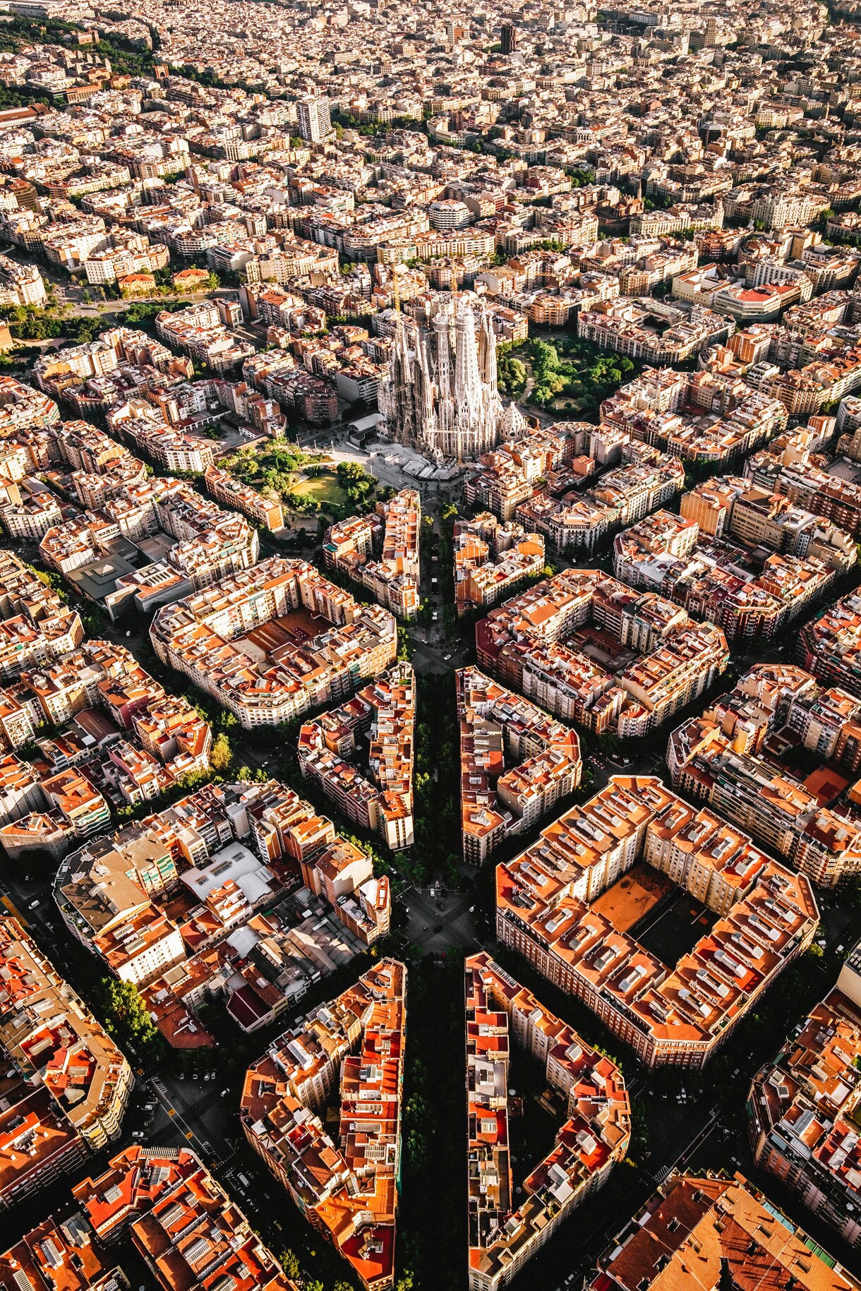 Aerial view of Barcelona centered on La Sagrada Família, surrounded by tightly packed city blocks with red rooftops and intersecting streets forming a geometric grid.