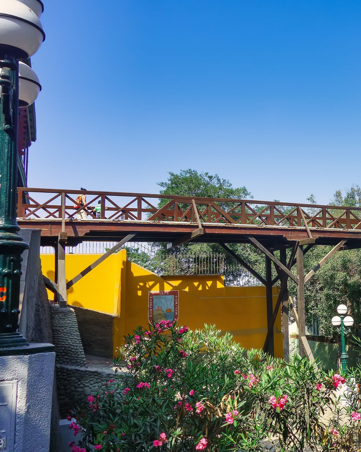 View of the wooden Bridge of Sighs in Barranco, Lima, surrounded by bright yellow buildings, palm trees and lush flowering plants under a clear blue sky.