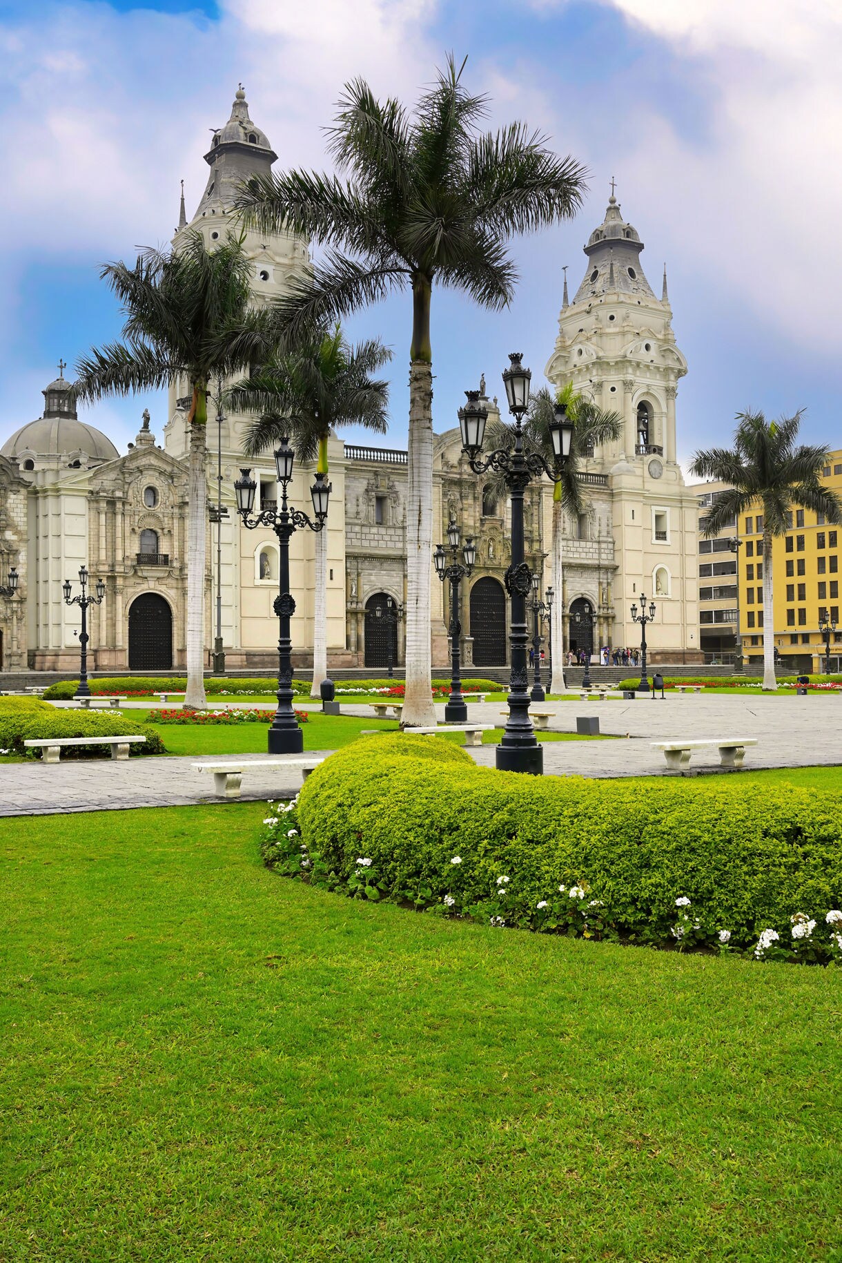 Lush green gardens with palm trees and ornate black lampposts in front of the white stone Cathedral of Lima on a bright day.